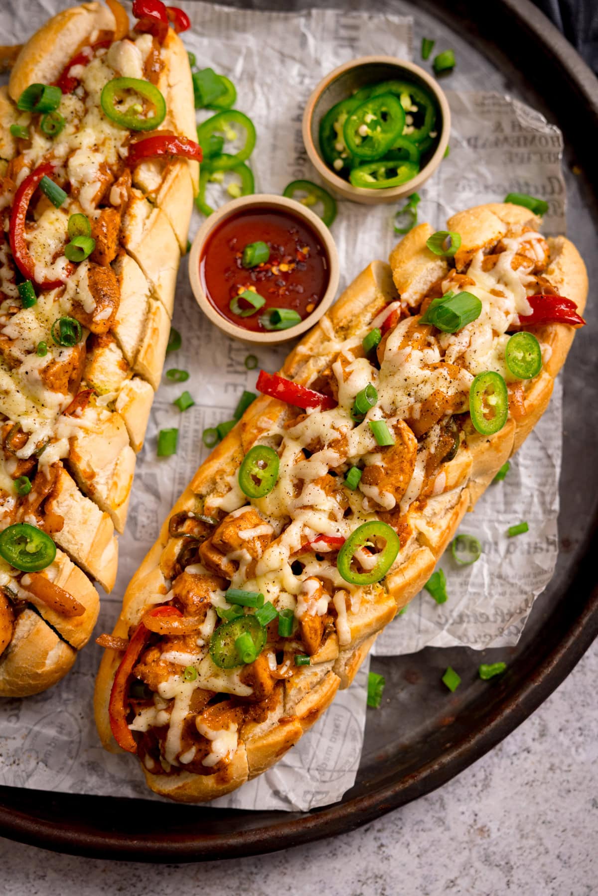 Two loaded Buffalo Chicken Garlic Bread sandwiches topped with melted cheese, sliced jalapeños, spring onions, and red peppers, served on a tray with small bowls of sauce and extra chopped spring onions.