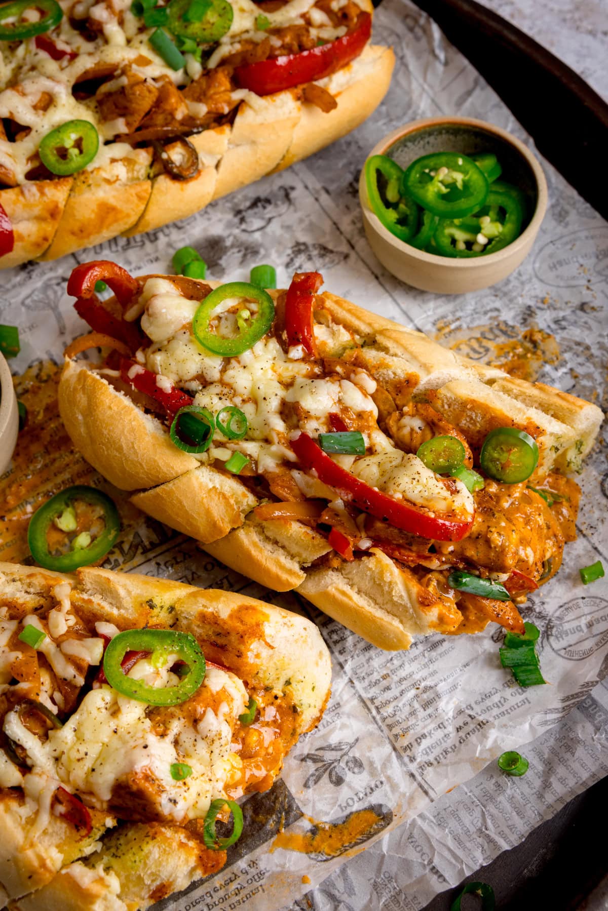 Half of a loaded Buffalo Chicken Garlic baguette topped with melted cheese, sliced jalapeños, red peppers, and spring onions on a tray lined with printed paper; a bowl of sliced jalapeños is nearby, along with 2 further stuffed baguettes in the edges of the shot.