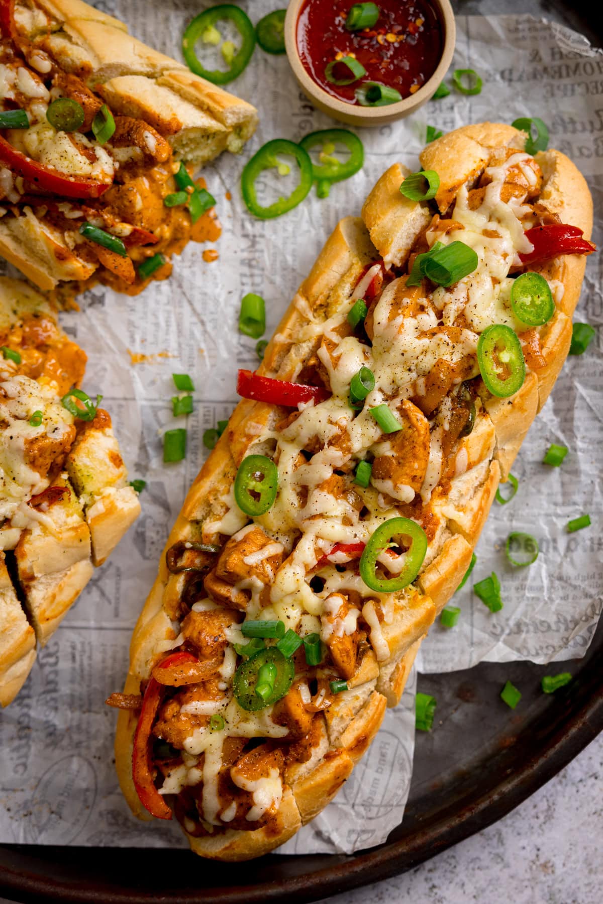 A overhead image of Buffalo chicken stuffed garlic bread filled with melted cheese, sliced jalapeños, red peppers, and spring onions on baking parchment, served with a small bowl of sauce nearby.
There is also a further stuffed baguette just in shot.