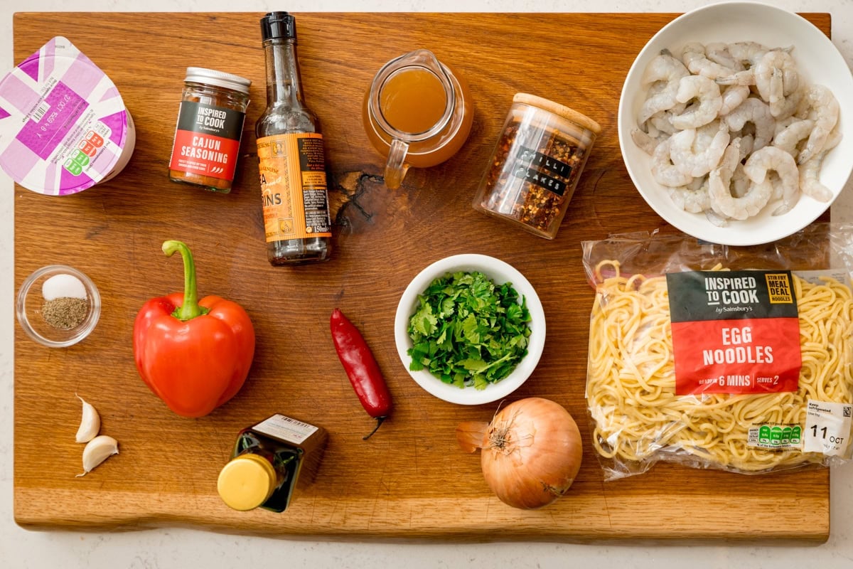 Ingredients for a Spicy Prawns with noodles on a wooden board. This includes: king prawns, egg noodles, red pepper, chilli, garlic, onion, coriander, seasonings and stock.