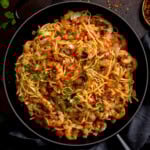 A frying pan filled with king prawn stir-fry noodles, featuring prawns, noodles, diced red peppers, onions, and garnished with chopped herbs.