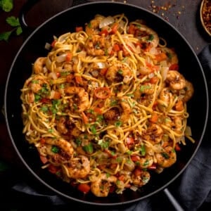 A frying pan filled with king prawn stir-fry noodles, featuring prawns, noodles, diced red peppers, onions, and garnished with chopped herbs.