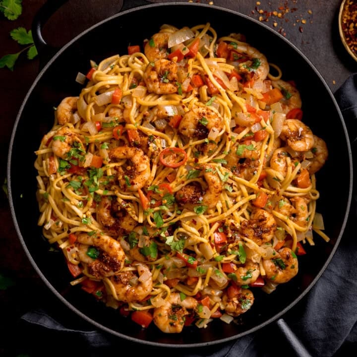A frying pan filled with king prawn stir-fry noodles, featuring prawns, noodles, diced red peppers, onions, and garnished with chopped herbs.
