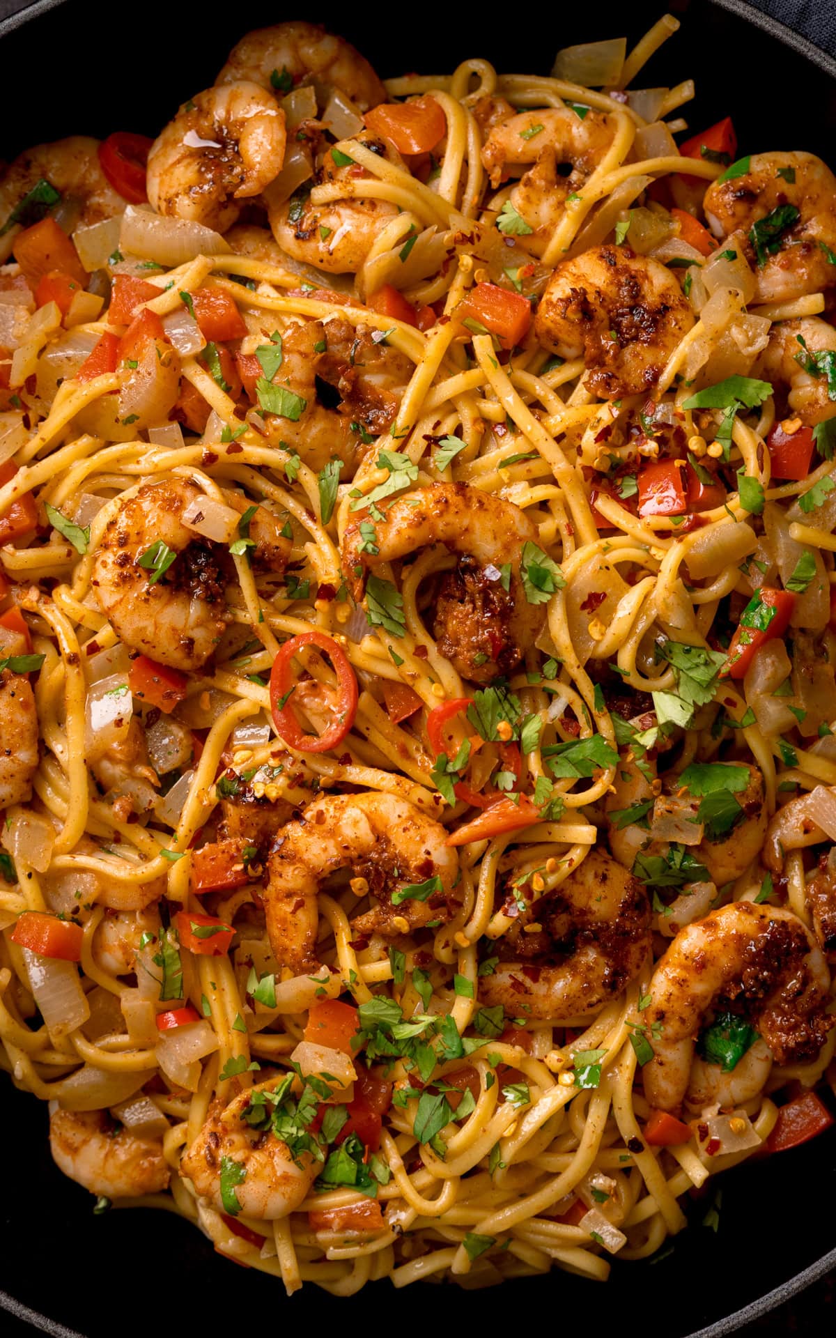 Close-up of a Cajun prawn noodle stir-fry with king prawns, red peppers, onions, and chopped coriander, garnished with chilli flakes.