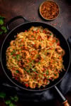 A frying pan filled with Cajun King Prawns with Noodles, garnished with chopped herbs, diced tomatoes, and onions, with a small bowl of crushed chilli flakes in the background.