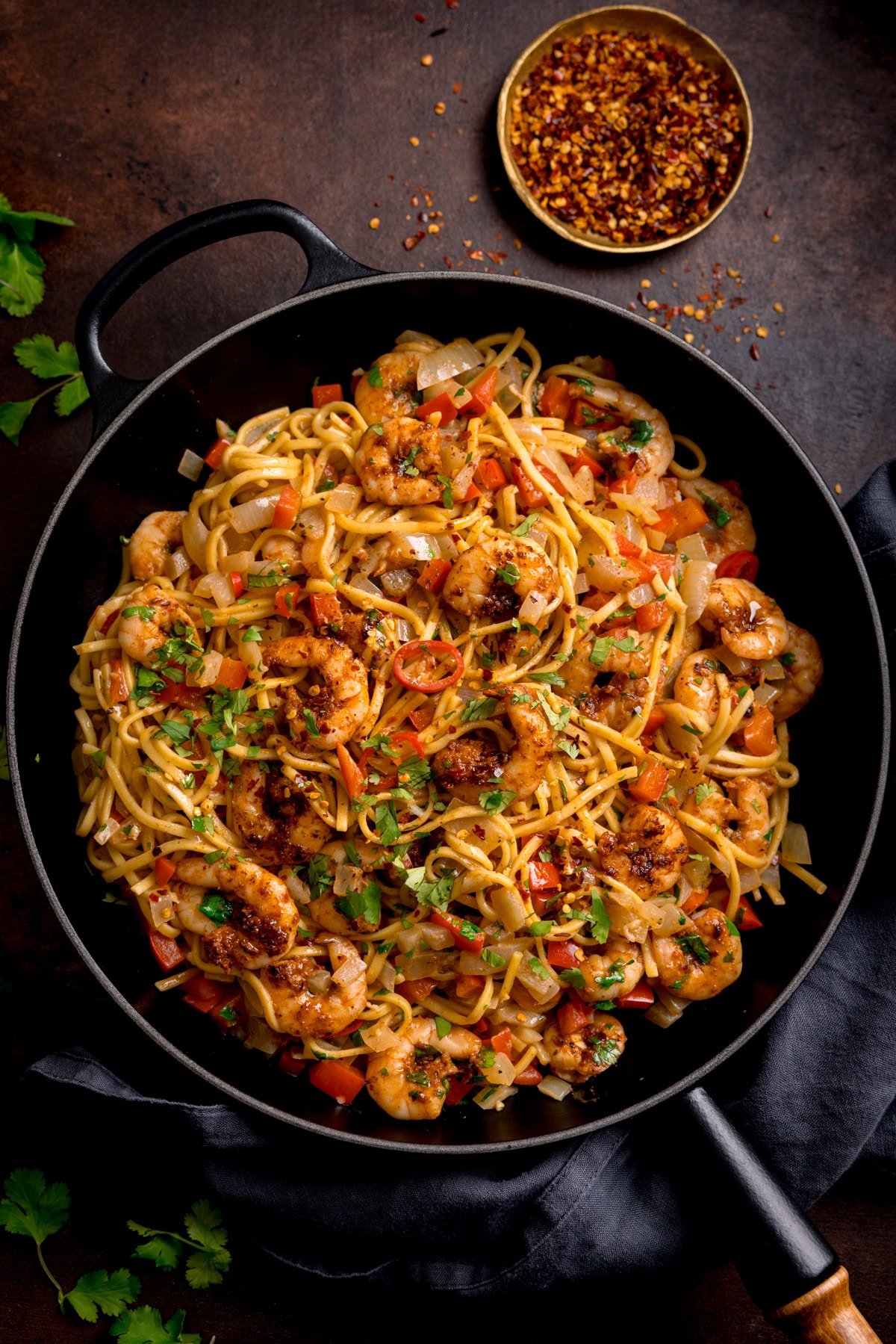 A frying pan filled with Cajun King Prawns with Noodles, garnished with chopped coriander. The pan is on a dark background and there is a small bowl of crushed chilli flakes in the background.