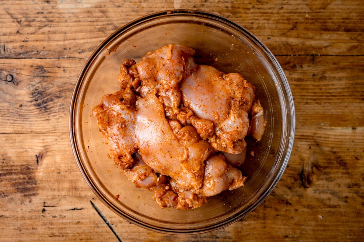 Raw chicken pieces coated with a seasoned marinade in a clear glass bowl, placed on a wooden surface.