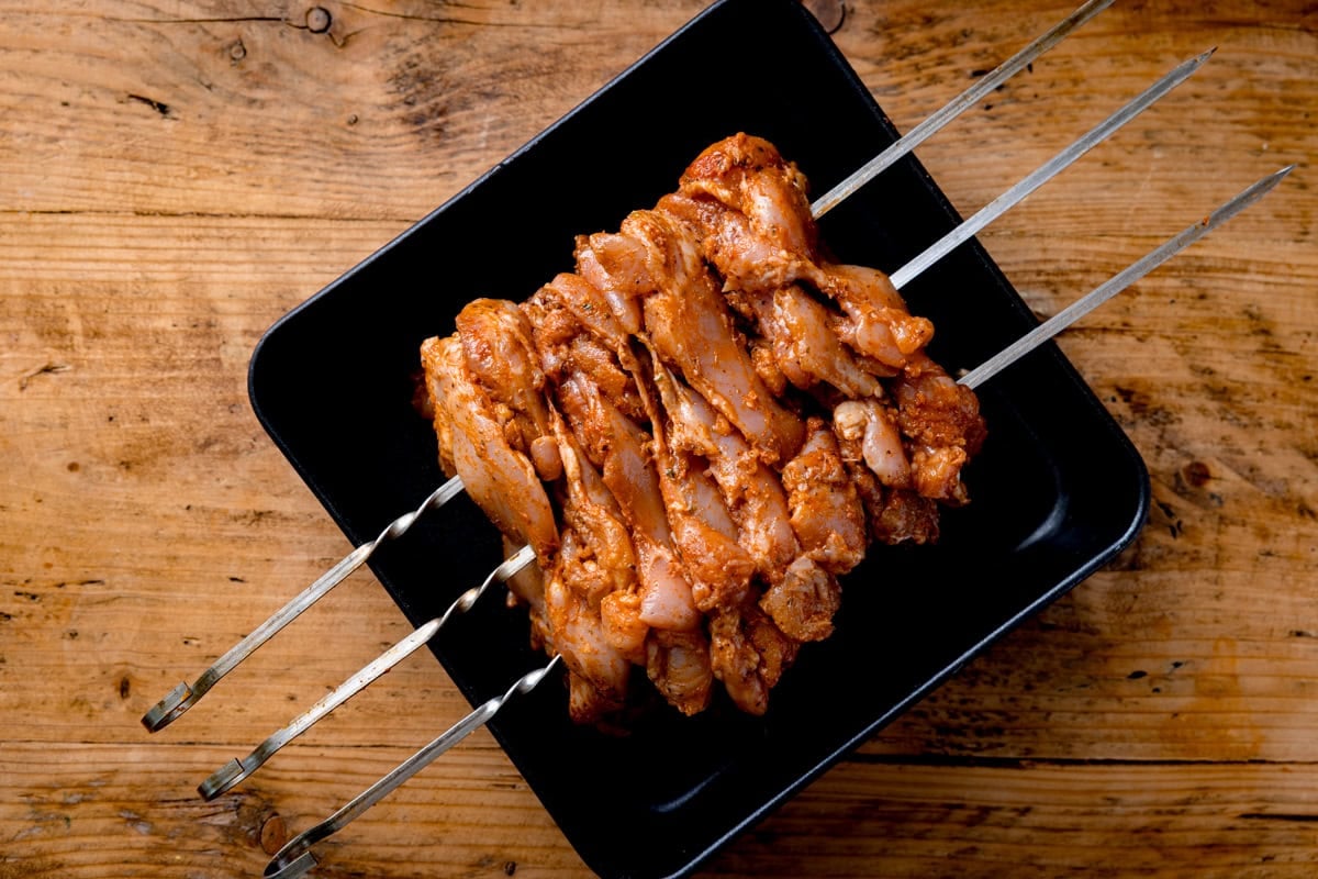 Raw marinated chicken pieces for a Chicken Doner Recipe are threaded onto three metal skewers and arranged on a black rectangular tray atop a wooden worktop.