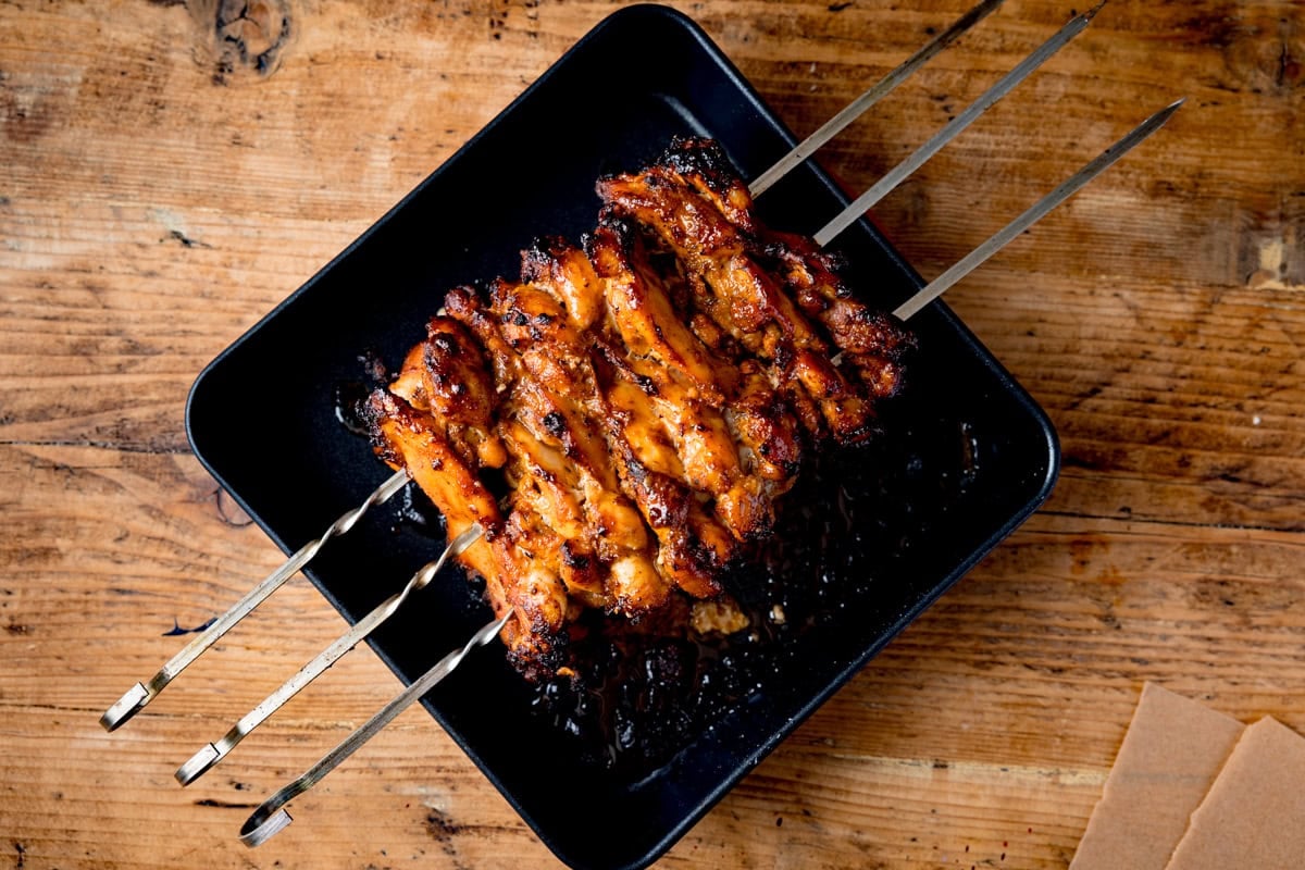 Grilled skewered chicken stacked on metal skewers, served in a black rectangular tray on a wooden surface.