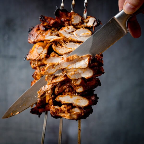 A close-up of a large knife slicing marinated, grilled chicken doner stacked on skewers.