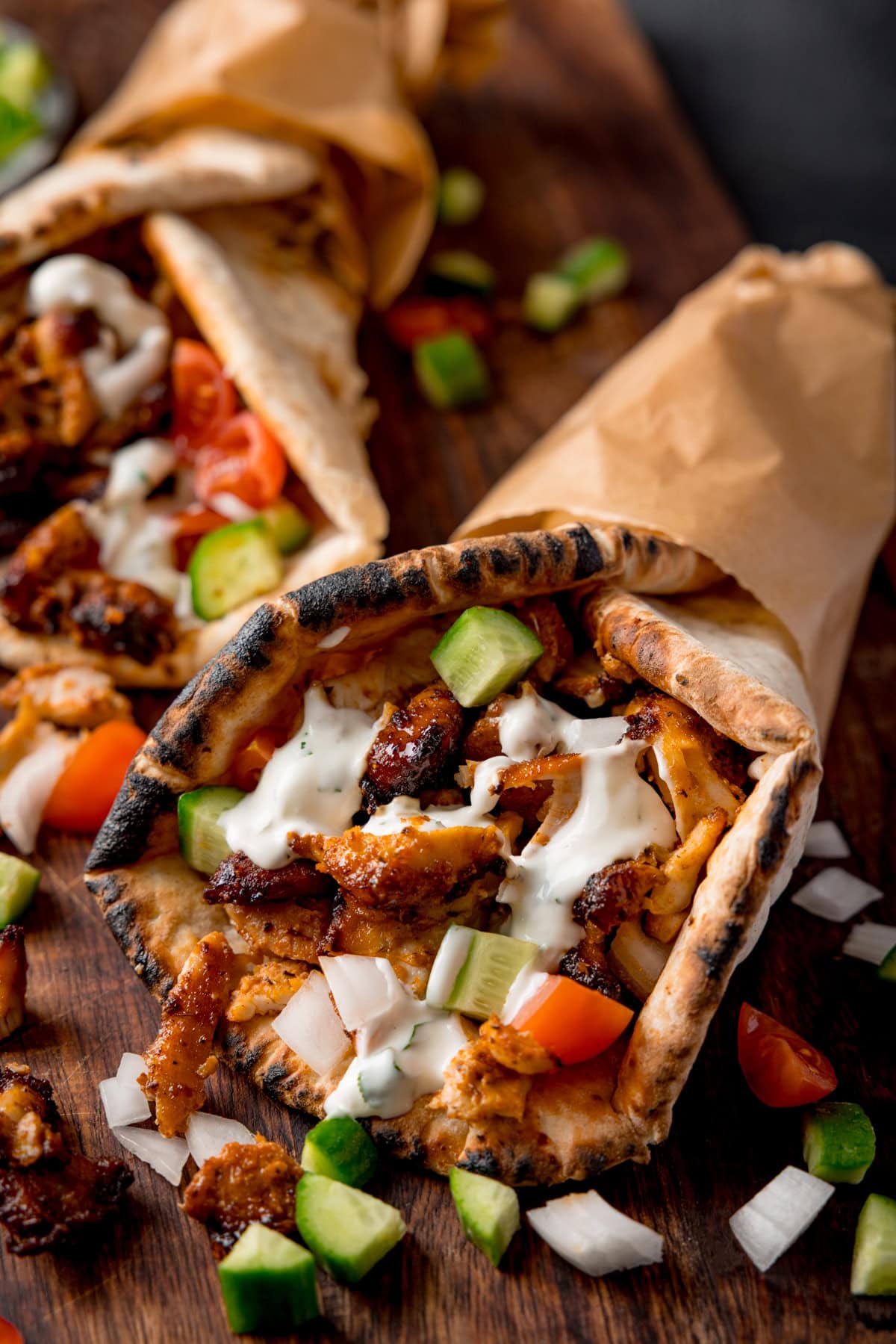 Two grilled chicken doners are wrapped in flatbreads with diced cucumber, tomato, onions, and garlic sauce, served on a wooden board.