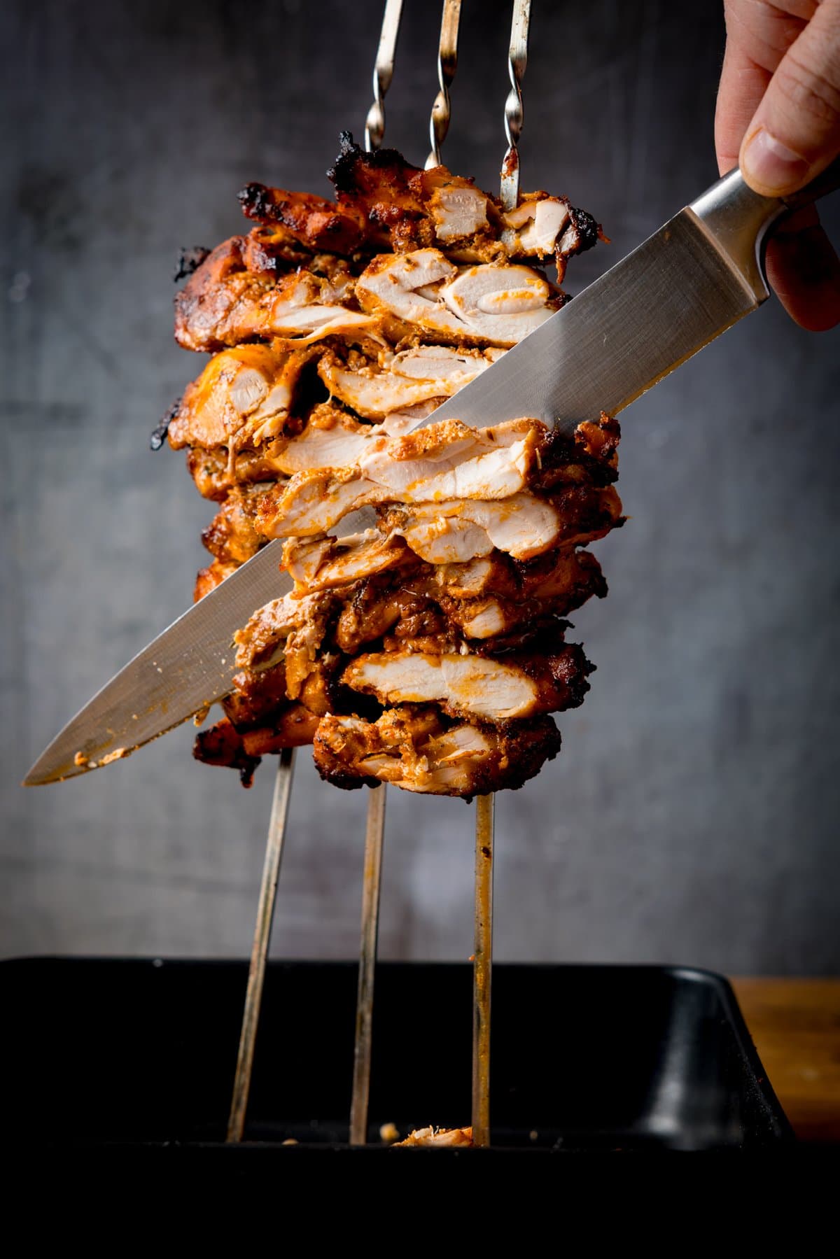 A hand holds a knife, slicing cooked, marinated chicken doner stacked on vertical skewers over a black tray.