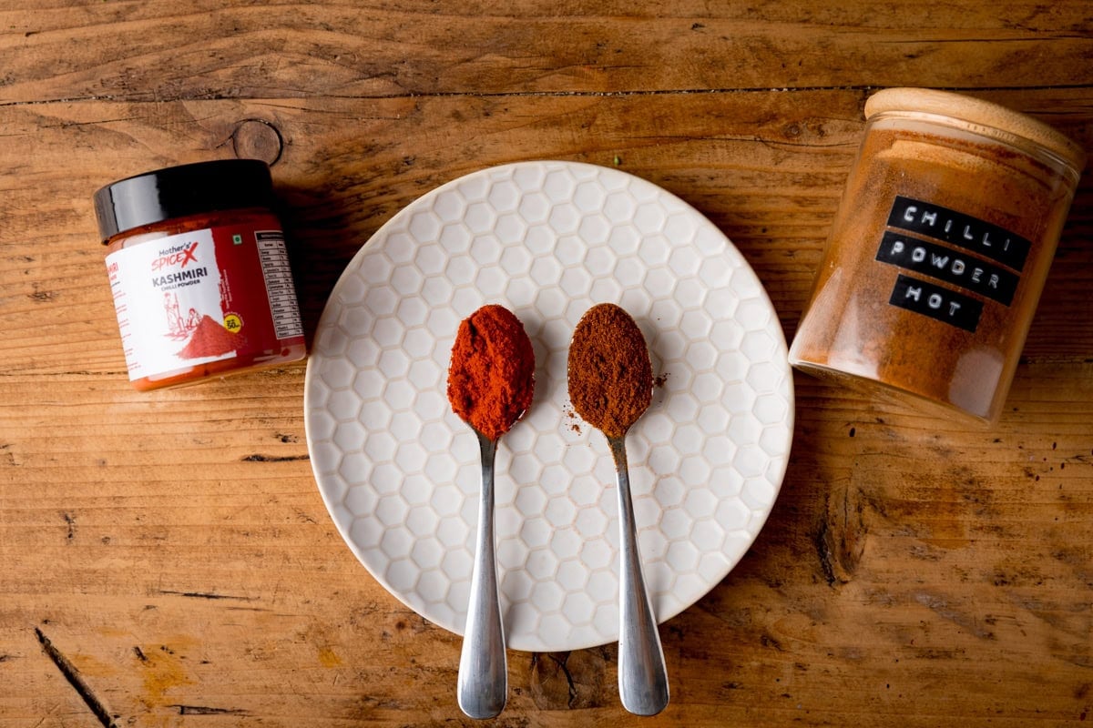 Two metal spoons with chilli powders on a white plate, next to a jar of Kashmiri chilli and a jar labelled chilli powder hot.