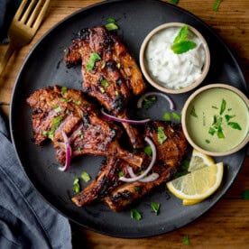 A black plate with succulent Tandoori Lamb Chops, sliced red onions, lemon wedges, green garnish, and two small bowls of dipping sauces on a wooden table.
