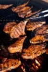 Tandoori Lamb Chops cooking over an open flame, with a pair of tongs lifting one perfectly grilled chop from the barbecue.