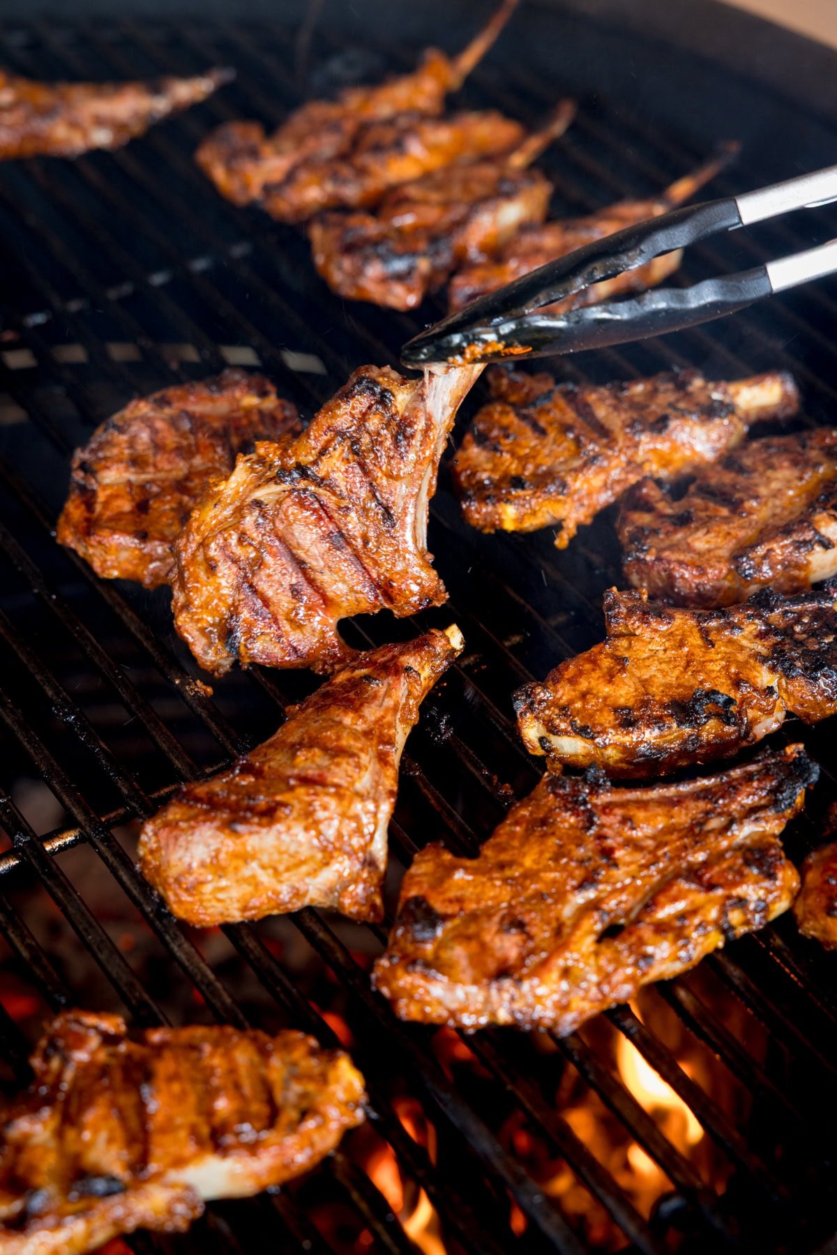 Tandoori Lamb Chops cooking over an open flame, with a pair of tongs lifting one grilled chop from the barbecue.