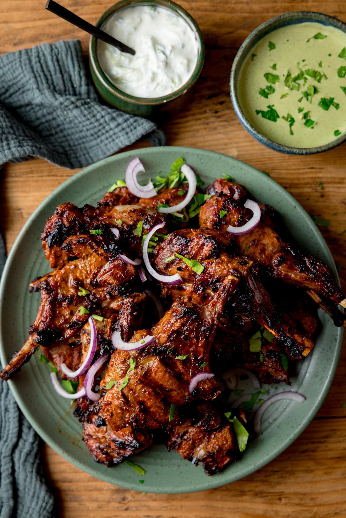 A green plate piled with grilled Tandoori Lamb Chops garnished with sliced red onions and coriander (cilantro), served with two dipping sauces in small bowls on a wooden table.