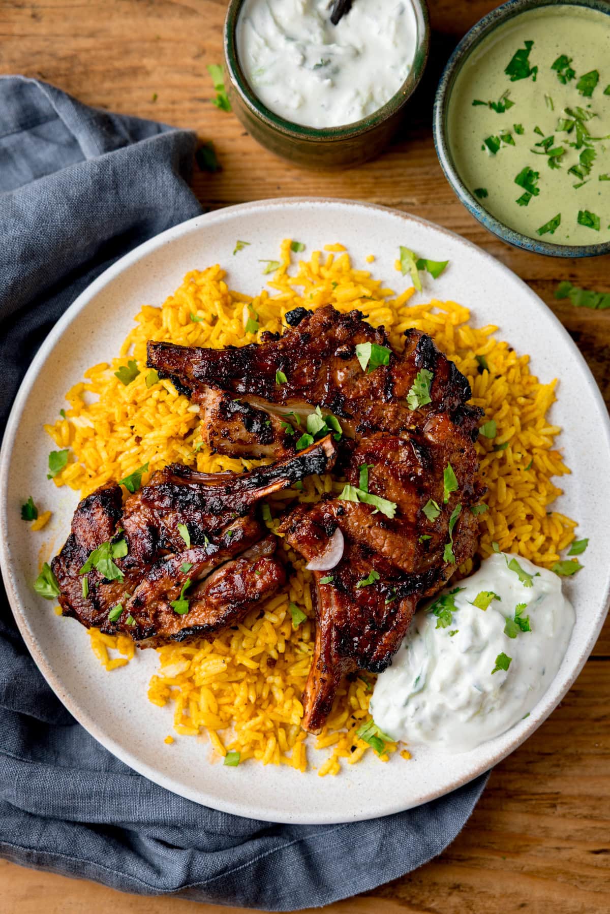 A white plate topped with Tandoori Lamb Chops on a bed of yellow rice with a dollop of raita, served alongside two bowls of sauces on a wooden table.