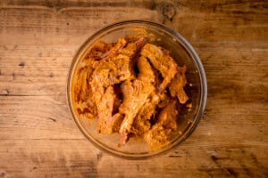 A glass bowl filled with Tandoori Lamb Chops marinating in a reddish-brown spice mixture sits on a rustic wooden surface.