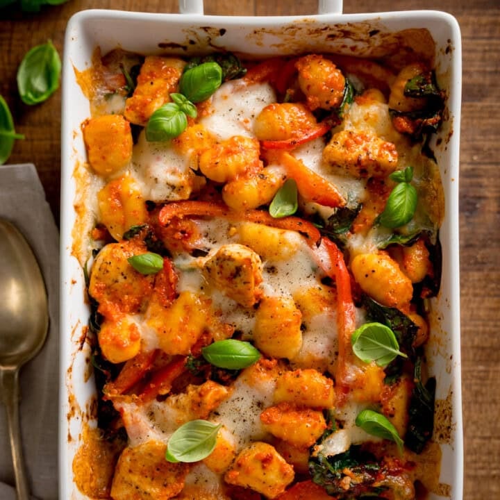 Overhead square image of baked gnocchi with melted cheese, tomatoes, spinach, and fresh basil in a white oven dish. The dish is on a wooden table with a spoon and napkin beside it.