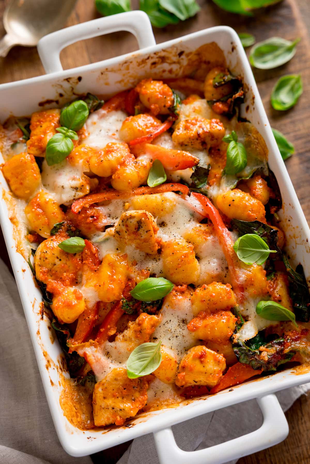 Overhead image of baked gnocchi with melted cheese, tomatoes, spinach, and fresh basil in a white oven dish. The dish is on a wooden table with a scattering of basil leaves next to it.