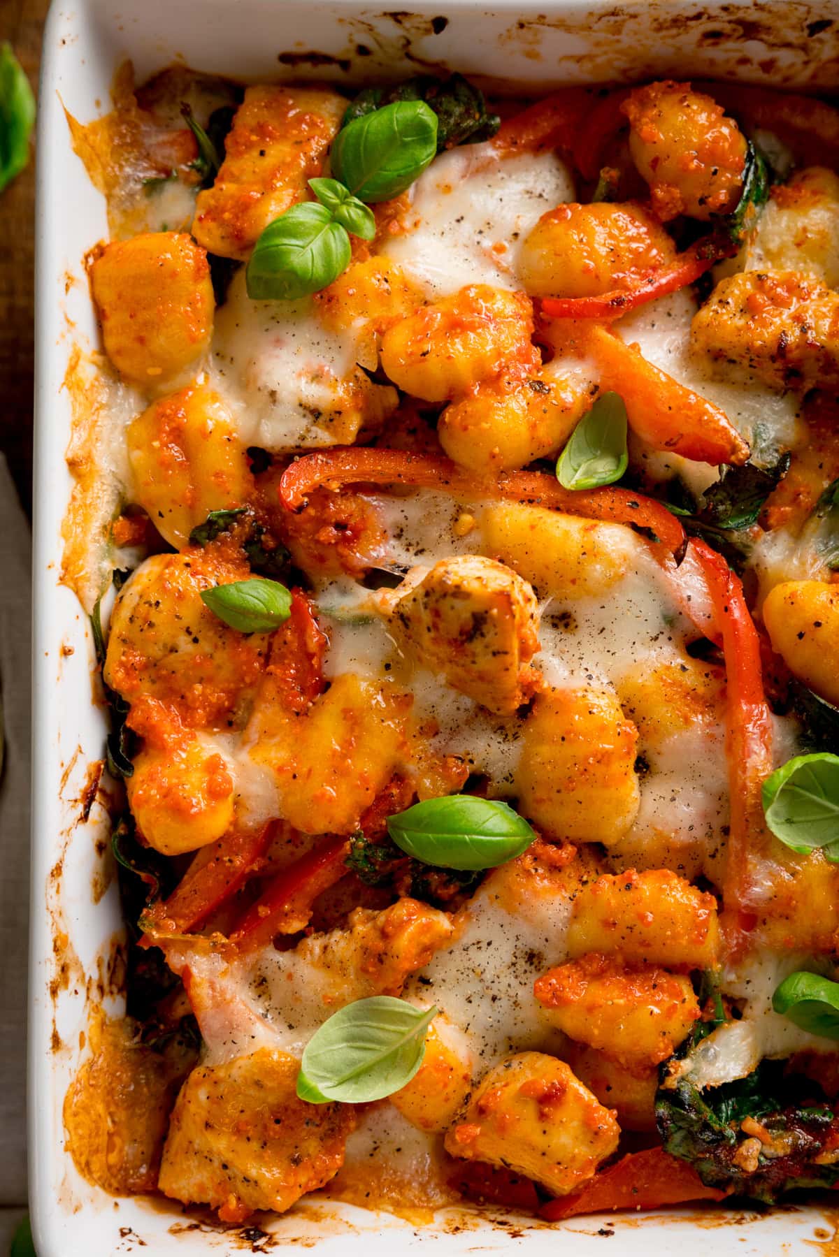 Overhead close up of baked gnocchi with melted cheese, tomatoes, spinach, and fresh basil in a white oven dish.