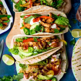 Three grilled chicken tacos with avocado, soured cream, jalapeños, coriander, and salsa on flour tortillas, served with lime wedges on a blue surface.