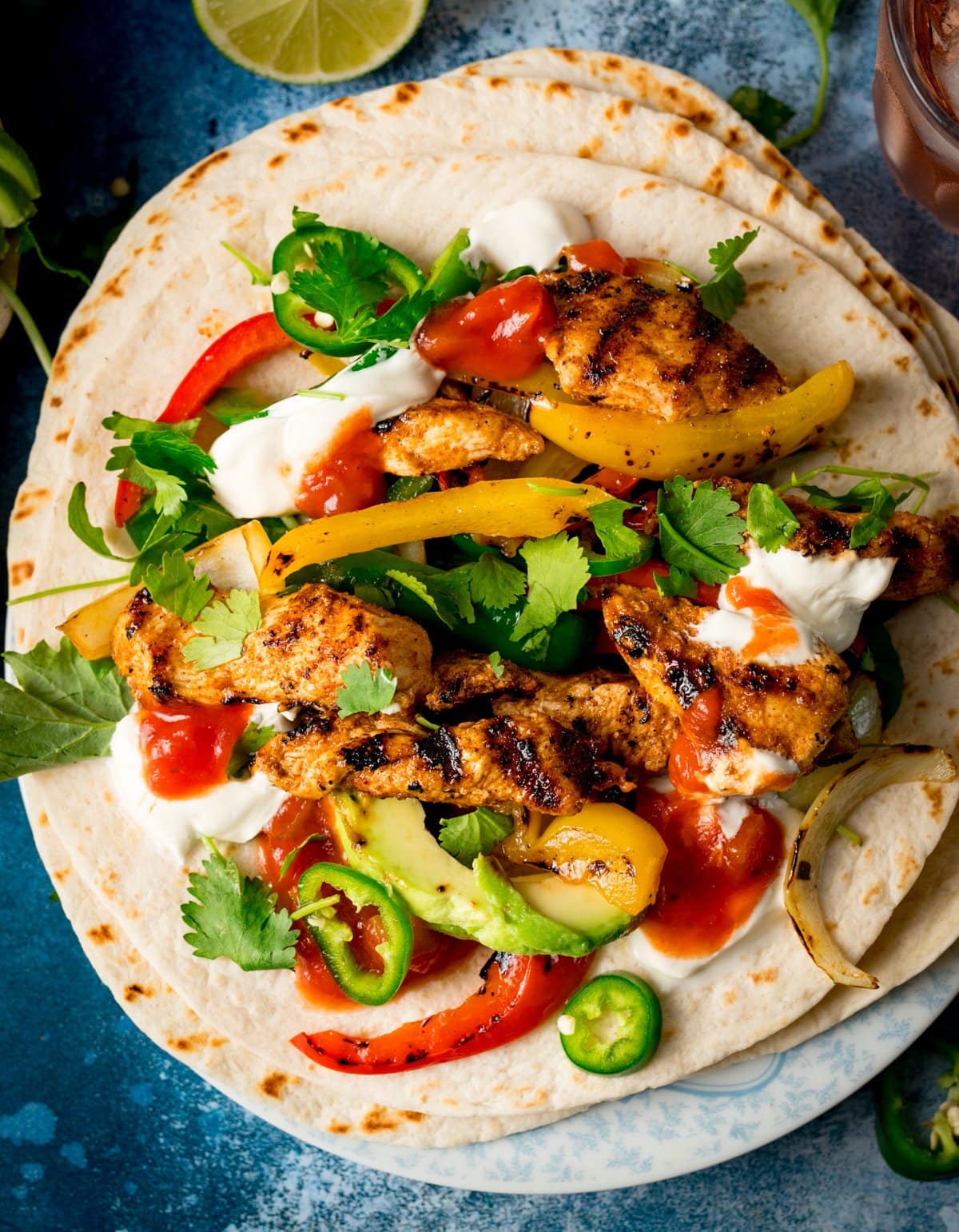 Two flour tortillas topped with grilled chicken, peppers, onions, avocado, jalapeño slices, coriander, salsa, and soured cream on a blue background.