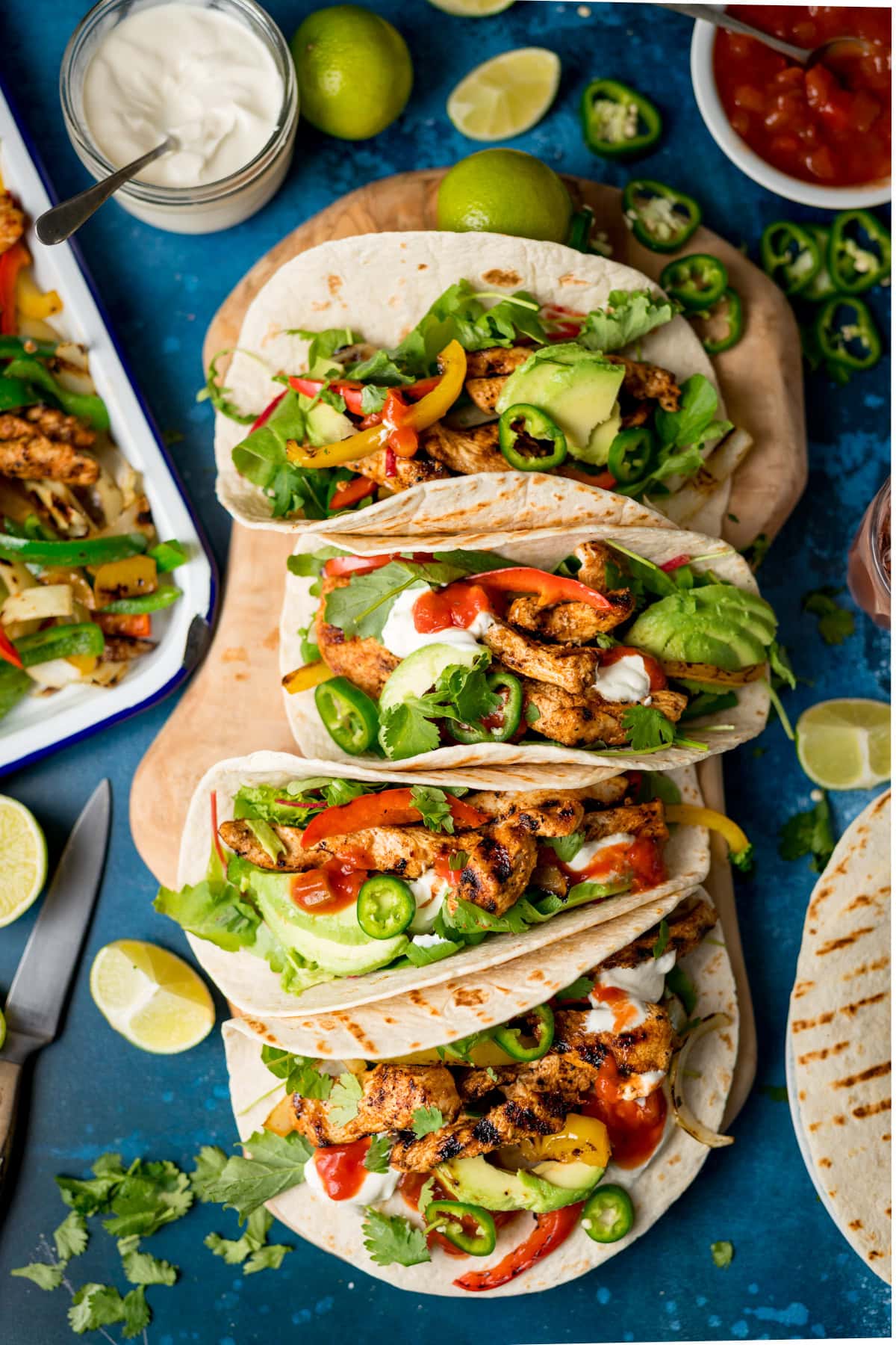 Three grilled chicken fajitas with peppers, onions, avocado, jalapeños, salsa and soured cream on tortillas, arranged on a blue surface with limes, salsa, and coriander nearby.