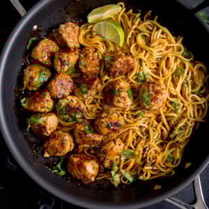 A frying pan containing cooked noodles and browned lemongrass meatballs, garnished with chopped herbs and lime wedges.