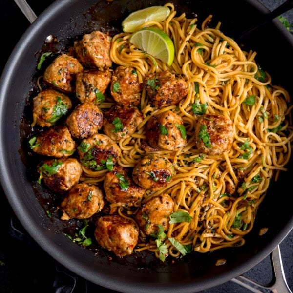 A frying pan containing cooked noodles and browned lemongrass meatballs, garnished with chopped herbs and lime wedges.