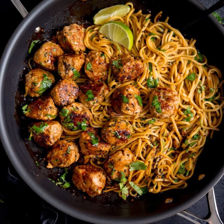 A frying pan containing cooked noodles and browned lemongrass meatballs, garnished with chopped herbs and lime wedges.