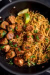 A frying pan filled with cooked noodles and pork meatballs, garnished with chopped coriander, lemongrass, and lime wedges.
