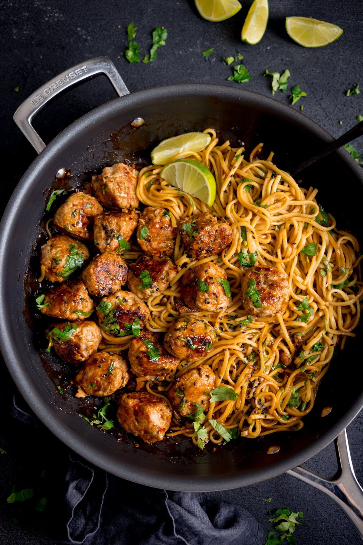 A frying pan containing cooked noodles and browned pork lemongrass meatballs, garnished with chopped cilantro and lime wedges. The pan is on a dark surface.