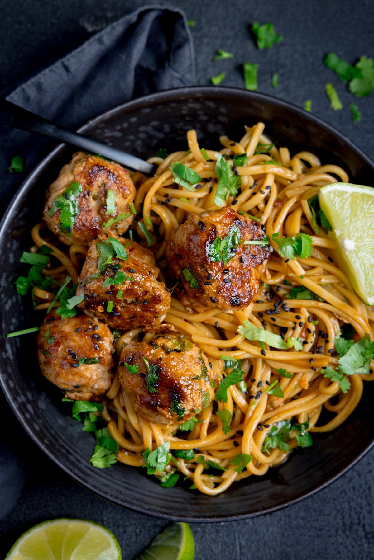 A black bowl filled with noodles, lemongrass meatballs, fresh coriander, black sesame seeds, and a lime wedge, with a fork resting at the side.