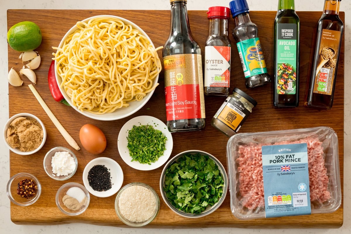 Ingredients for pork and lemongrass meatballs with noodles on a wooden board. Including egg noodles, pork mince, sauces, chopped herbs, brown sugar, egg, lime, garlic, and small bowls of spices and seasonings.