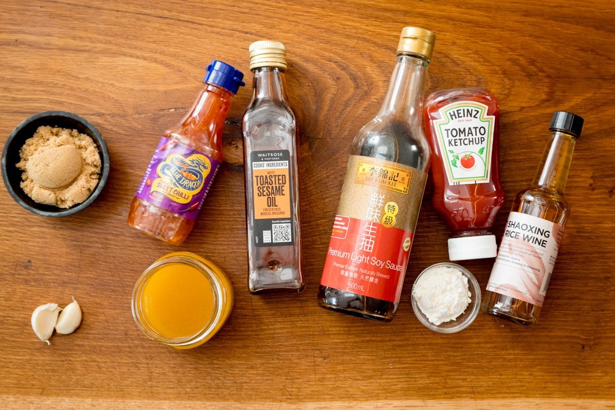 Various sauce ingredients for a homemade stir-fry sauce are arranged on a wooden surface, including brown sugar, sriracha, sesame oil, soy sauce, tomato ketchup, Shaoxing wine, cornflour, honey, and garlic cloves.