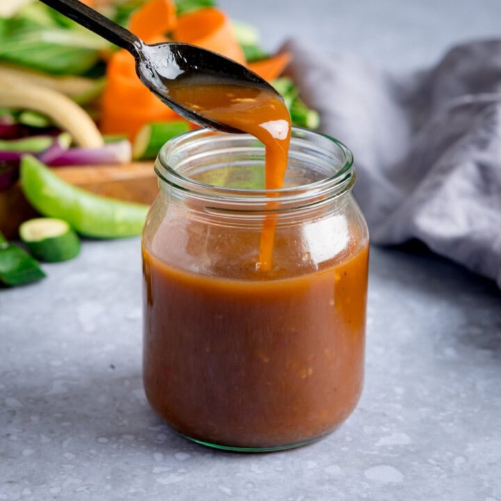 A glass jar filled with homemade stir fry sauce sits on a grey surface. A spoon above the jar drips sauce back inside. Fresh vegetables are slightly out of focus in the background, ideal for any stir fry sauce recipe.