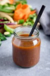 A glass jar filled with homemade stir-fry sauce and a black spoon, with assorted vegetables blurred in the background on a grey surface.