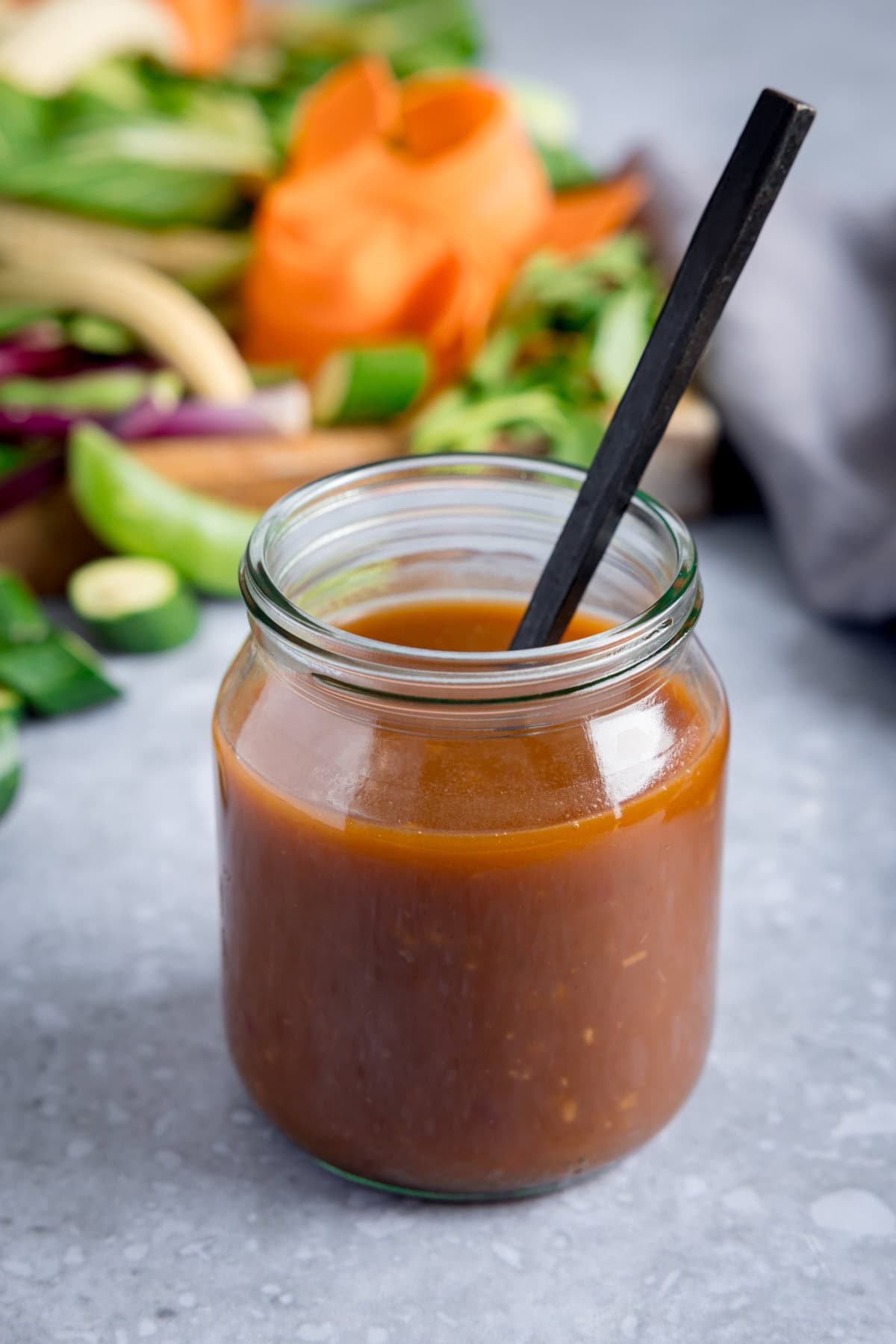 A glass jar filled with homemade stir-fry sauce and a black spoon, with assorted vegetables blurred in the background on a grey surface.