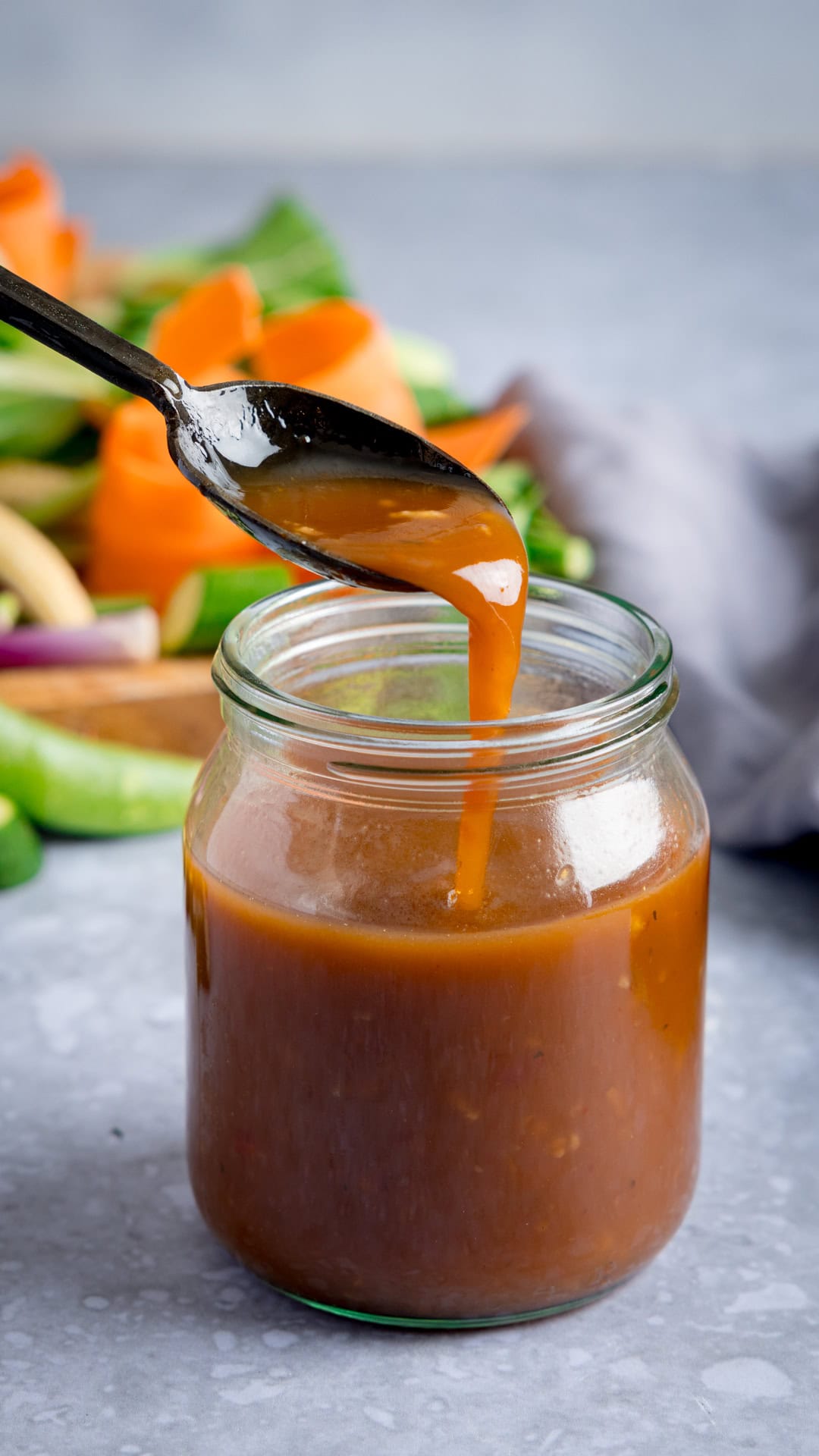 A spoon drizzles homemade stir-fry sauce back into a glass jar, with vegetables and a grey cloth in the blurred background.