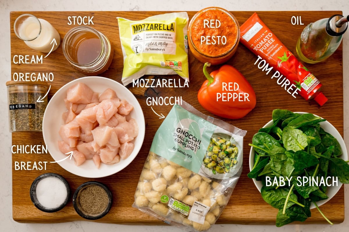 A wooden board with ingredients for baked gnocchi with chicken: diced chicken, gnocchi, mozzarella, cream, oregano, chicken stock, red pesto, red pepper, tomato purée, oil, baby spinach, salt and pepper.
The ingredients are labelled in white text.