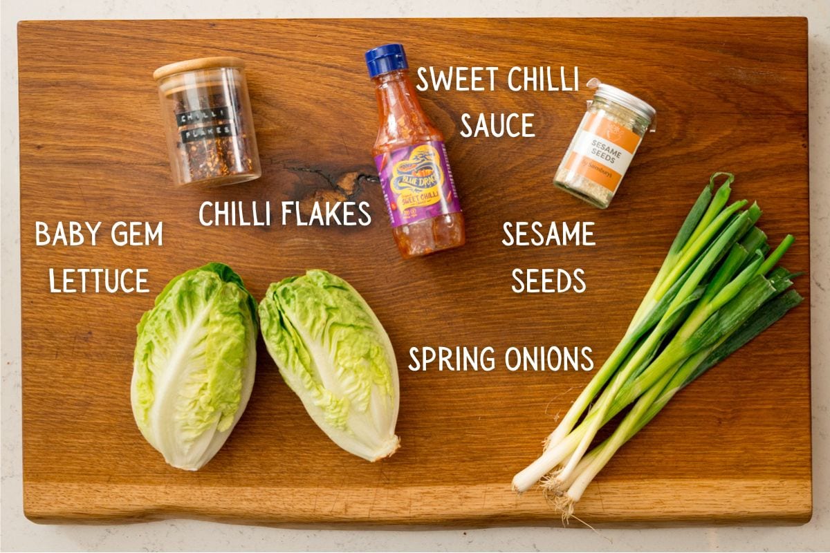 A wooden board with baby gem lettuce, spring onions, a bottle of sweet chilli sauce, a jar of chilli flakes, and a jar of sesame seeds.