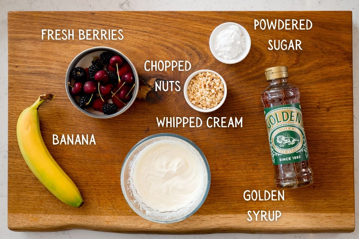 A wooden board displaying ingredients for French Toast toppings: a banana, a bowl of fresh berries, chopped nuts, whipped cream, icing sugar, and golden syrup, all labelled.
