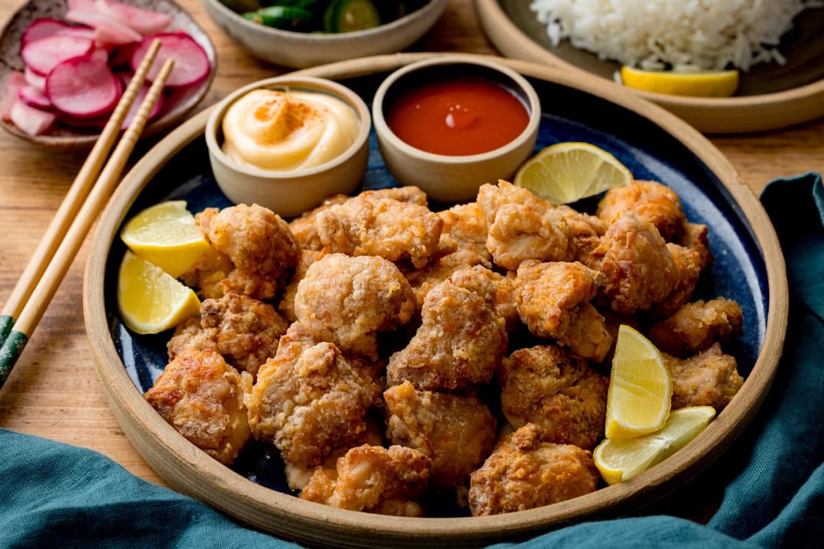 A plate of Air Fryer Chicken Karaage with lemon wedges, two dipping sauces, chopsticks, pickled vegetables, and a side of white rice in the background.
