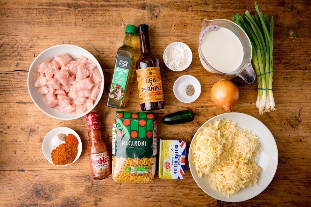Top-down view of ingredients for an easy buffalo chicken mac and cheese: chicken pieces, grated cheese, macaroni, spices, hot sauce, oil, Worcestershire sauce, flour, milk, butter, jalapeño, spring onions and onion on a wooden table.