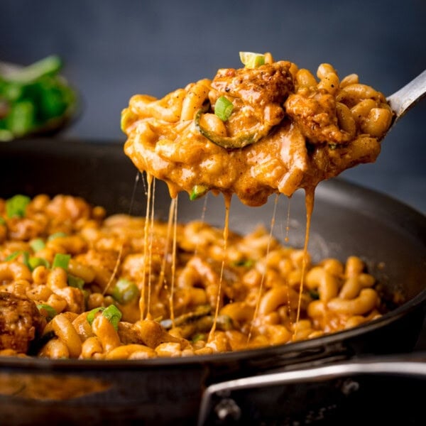 A spoon lifts cheesy Buffalo Chicken Macaroni Cheese with tender chicken and spring onions from a frying pan filled with creamy pasta.