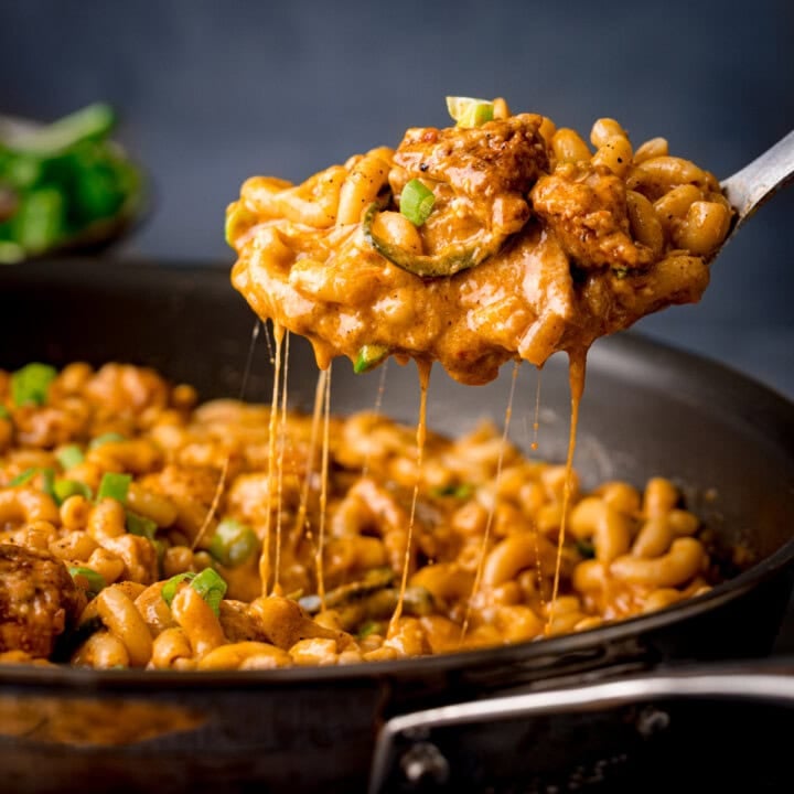 A spoon lifts cheesy Buffalo Chicken Macaroni Cheese with tender chicken and spring onions from a frying pan filled with creamy pasta.