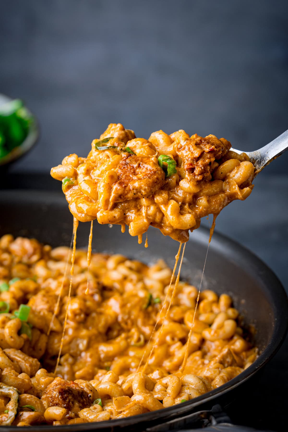 A spoon lifts cheesy Buffalo Chicken Macaroni Cheese mixed with chunks of chicken and spring onions from a pan, with melted cheese stretching between the spoon and the pan.