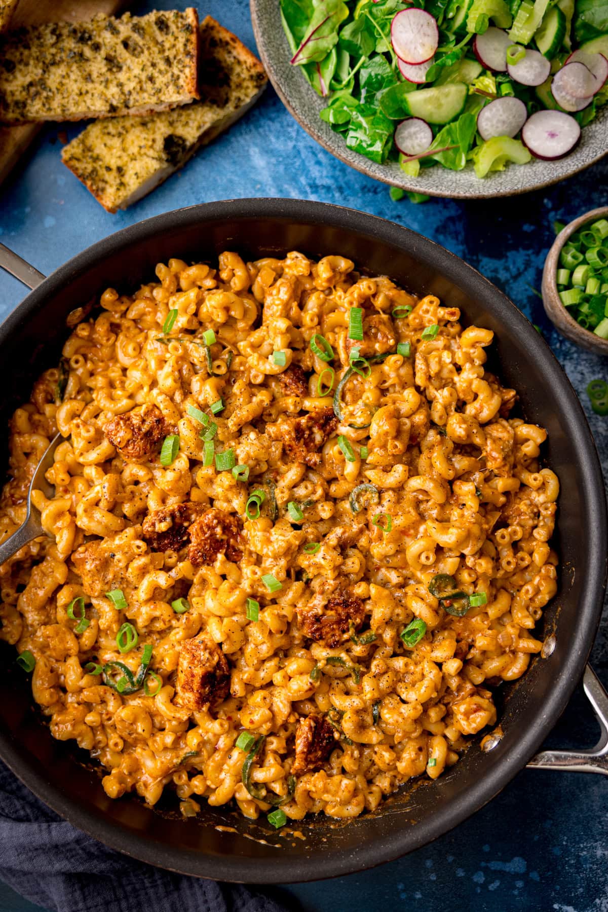 A frying pan filled with creamy Buffalo Chicken Macaroni Cheese, topped with pieces of sausage and chopped spring onions, served with salad and bread on the side.
